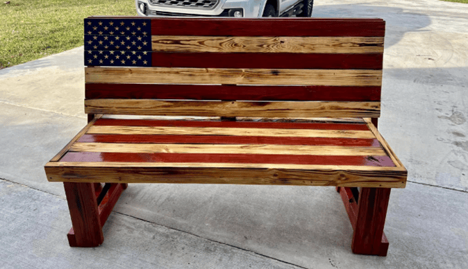 A handmade wooden bench painted like the American flag, photographed outdoors on a driveway.